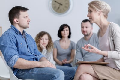 a man sitting in group therapy and listening to the psychologist with concerned family in the back